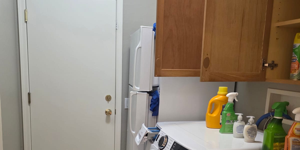 Laundry room interior door with a brass deadbolt beside a stacked washer and dryer, with cleaning bottles on the counter