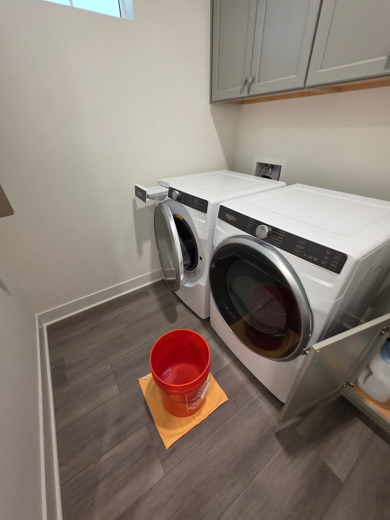 Laundry room with a front loading washer and dryer, gray upper cabinets and an orange bucket on a mat