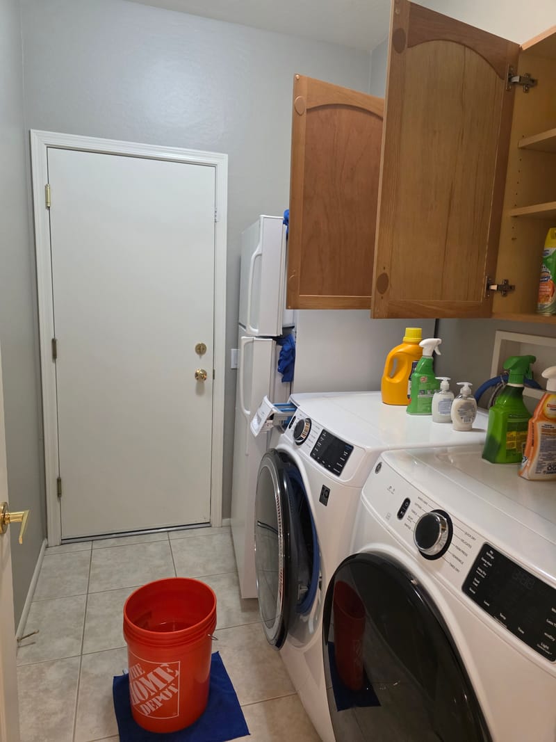 Laundry room with a front loading washer and dryer, wood cabinets above, a white door and an orange bucket on a blue mat on a tile floor