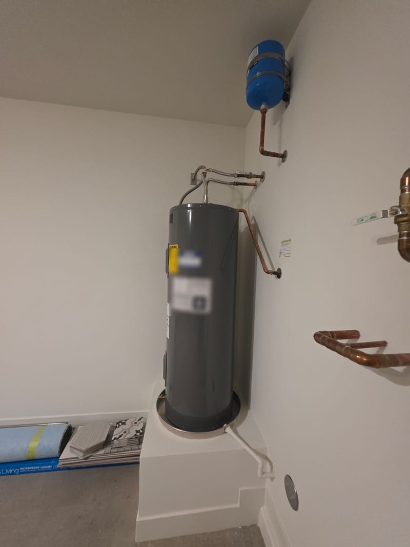 Electric water heater on a raised platform with a drain pan, copper supply lines and a blue expansion tank on the wall of a utility room